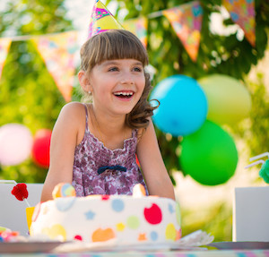 Little girl smiling, sitting in front of a cake with a birthday hat on, and balloons in the background