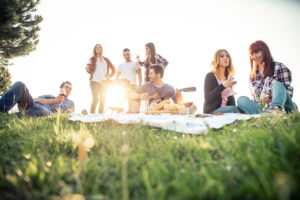 Group of friends sitting in the grass having a picnic, with one friend playing guitar while the others sip wine and smile
